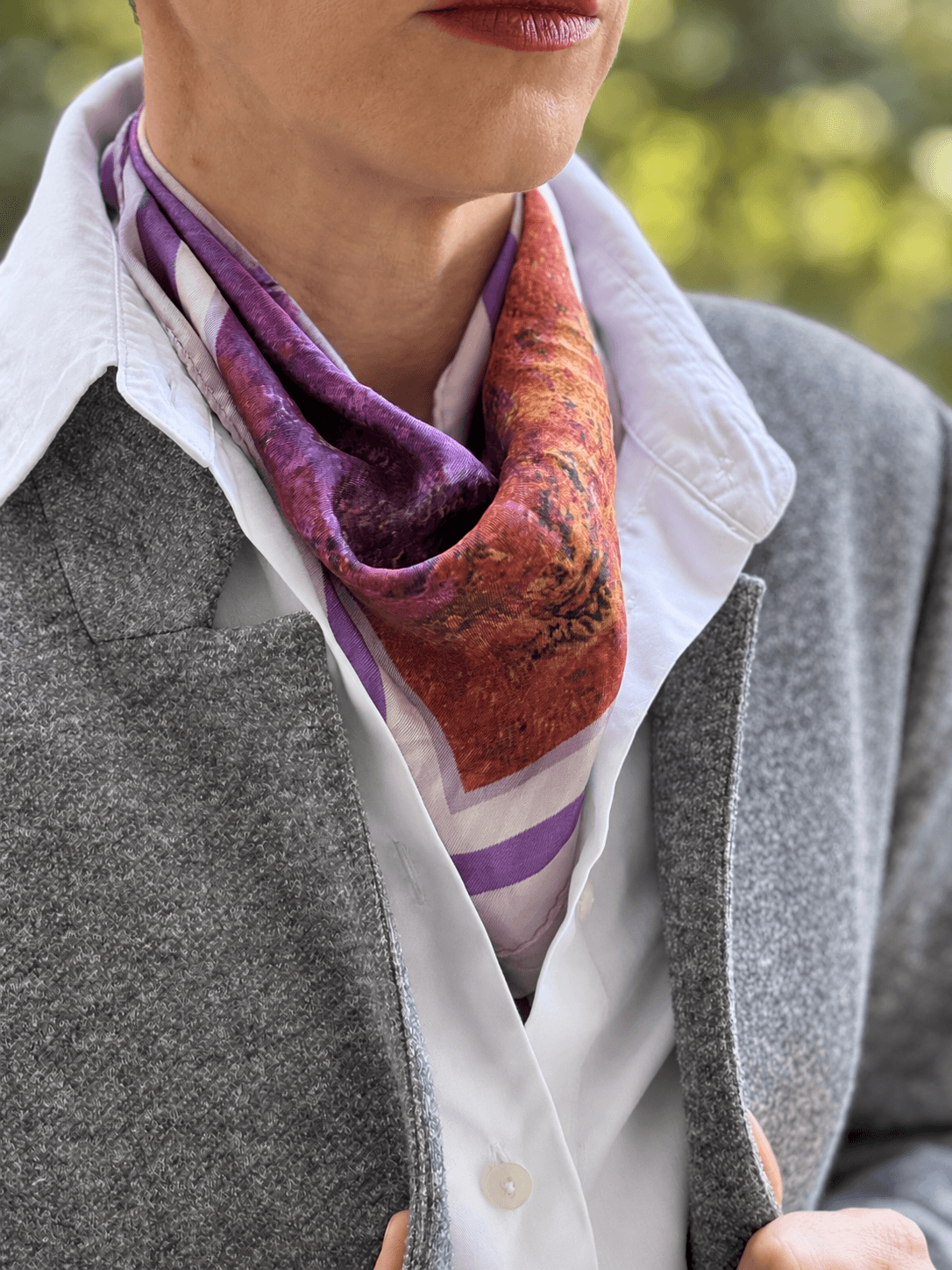 A close-up of a person wearing a patterned silk scarf in purple and rust orange, styled with a white collared shirt and a gray blazer.