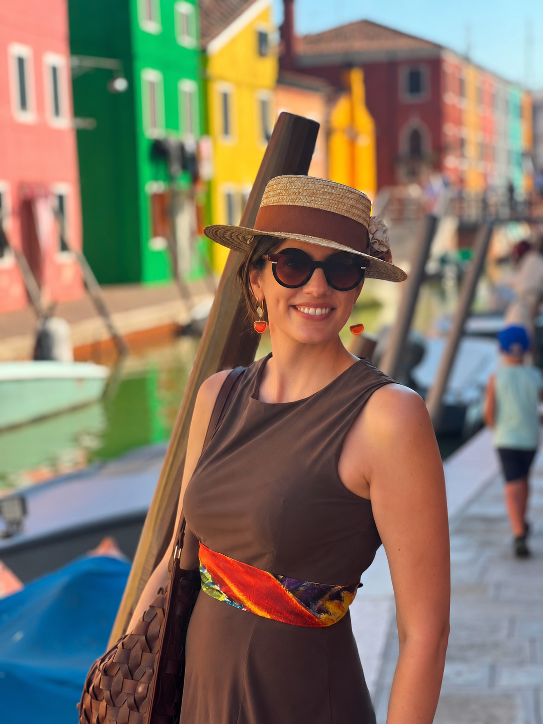 A woman wearing a brown sleeveless dress, a straw hat, a silk scarf and sunglasses stands on a canal boardwalk with colorful houses in the background.