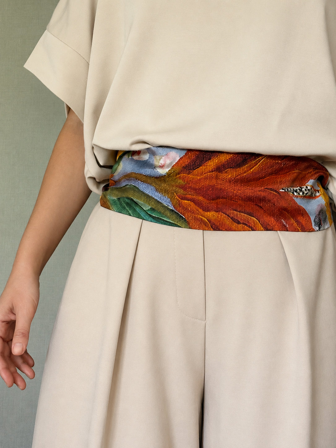 A close-up of a person wearing a cream-colored top and pleated trousers, tied at the waist with a wide, colorful abstract-patterned scarf in orange, red, green, and blue.