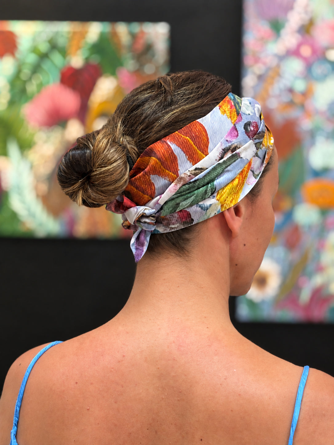A woman shown from the back wearing a colorful floral silk headband tied in a bun, with abstract paintings blurred in the background.