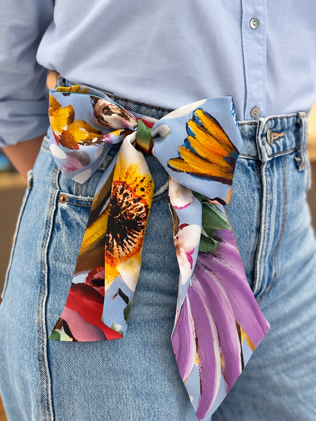 A close-up of a person wearing a light blue button-up shirt and blue jeans, with a colorful floral print fabric tie belt tied around the waist.