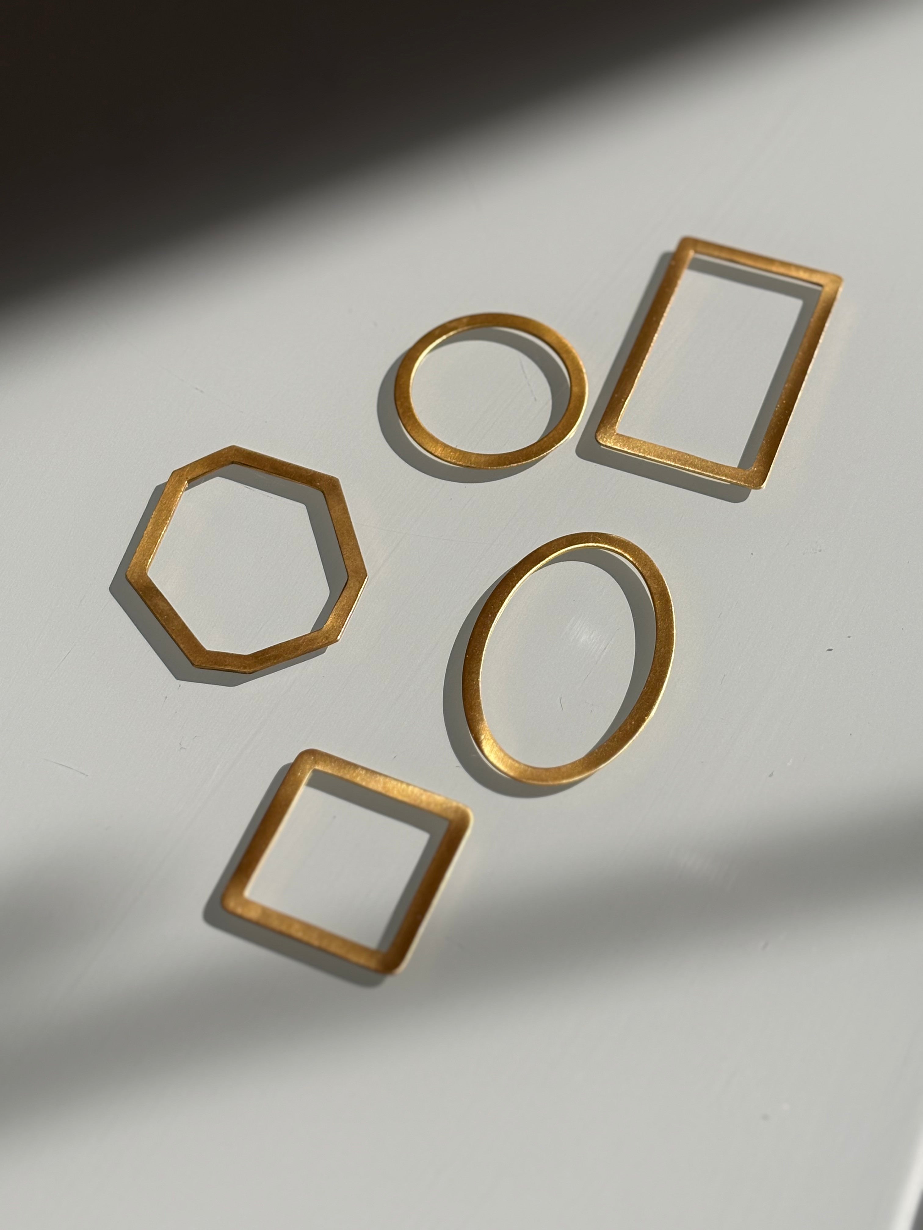 Five gold-toned metal geometric frames, including a hexagon, circle, two rectangles, and an oval, arranged on a white surface with soft shadows.