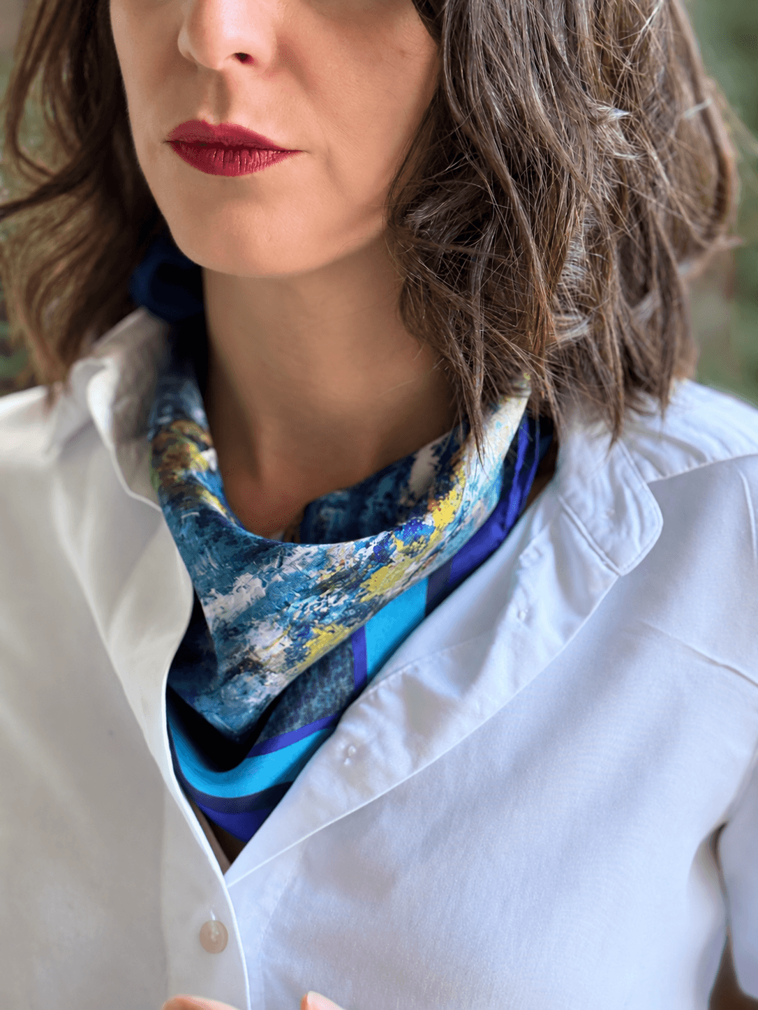 A close-up of a woman wearing a white button-up shirt and a patterned silk scarf in shades of blue, yellow, and green.
