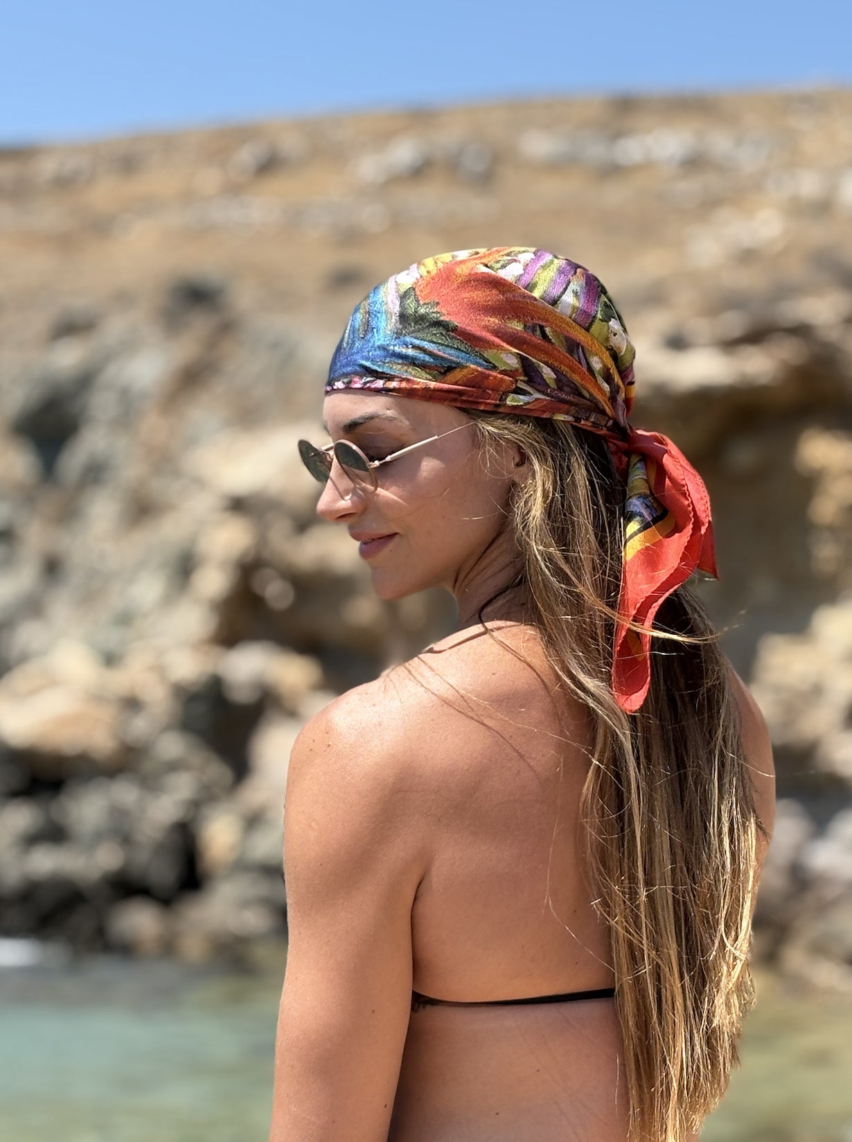 A woman in a black bikini top and sunglasses is shown from the back and side profile, wearing a vibrant, multicolored tropical print headscarf with a red side tie, set against a rocky beach background.