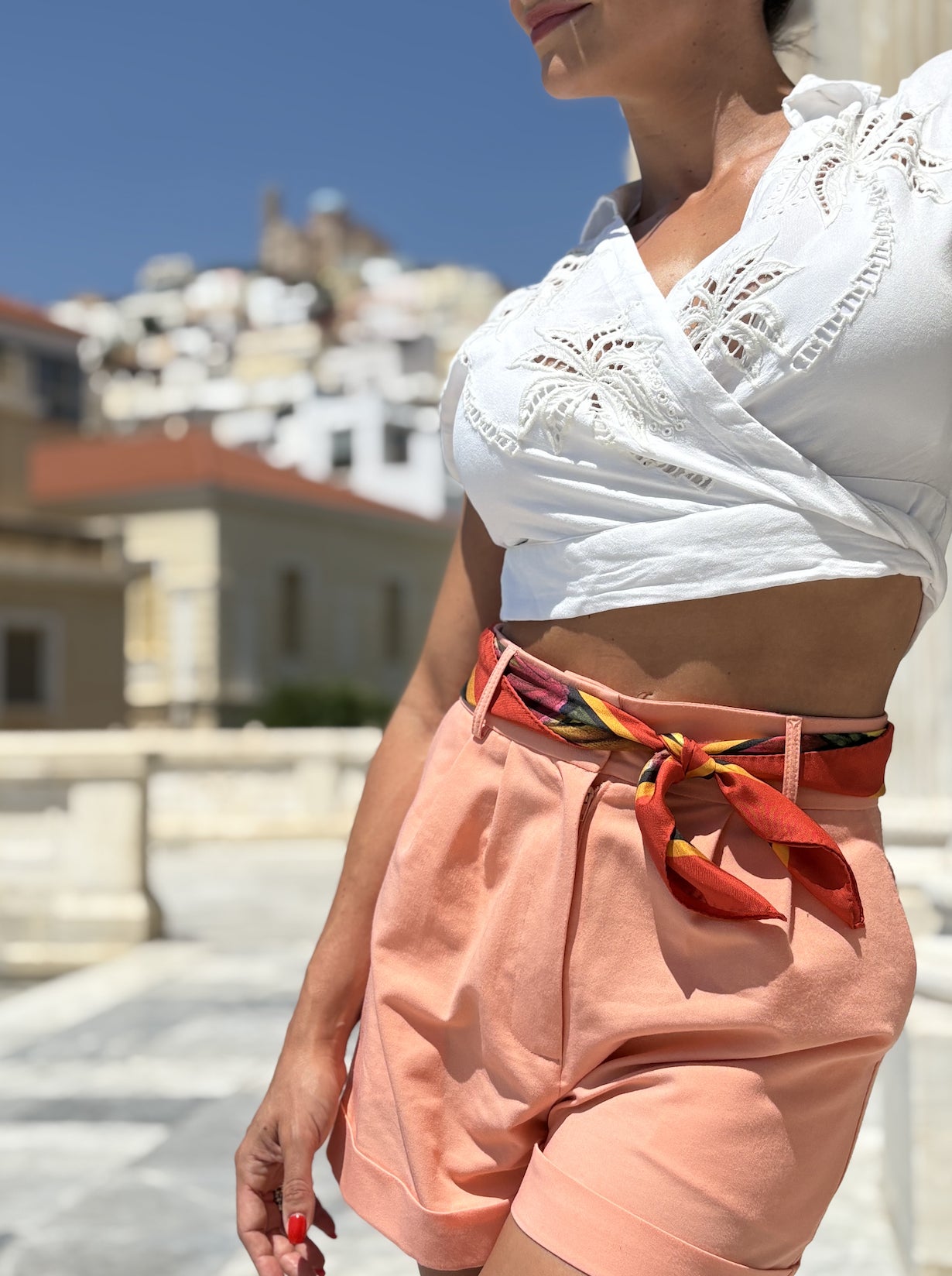 A woman wearing a white eyelet crop top and peach-colored shorts with a colorful patterned tie belt, photographed outdoors against a blurred background of buildings and blue sky.