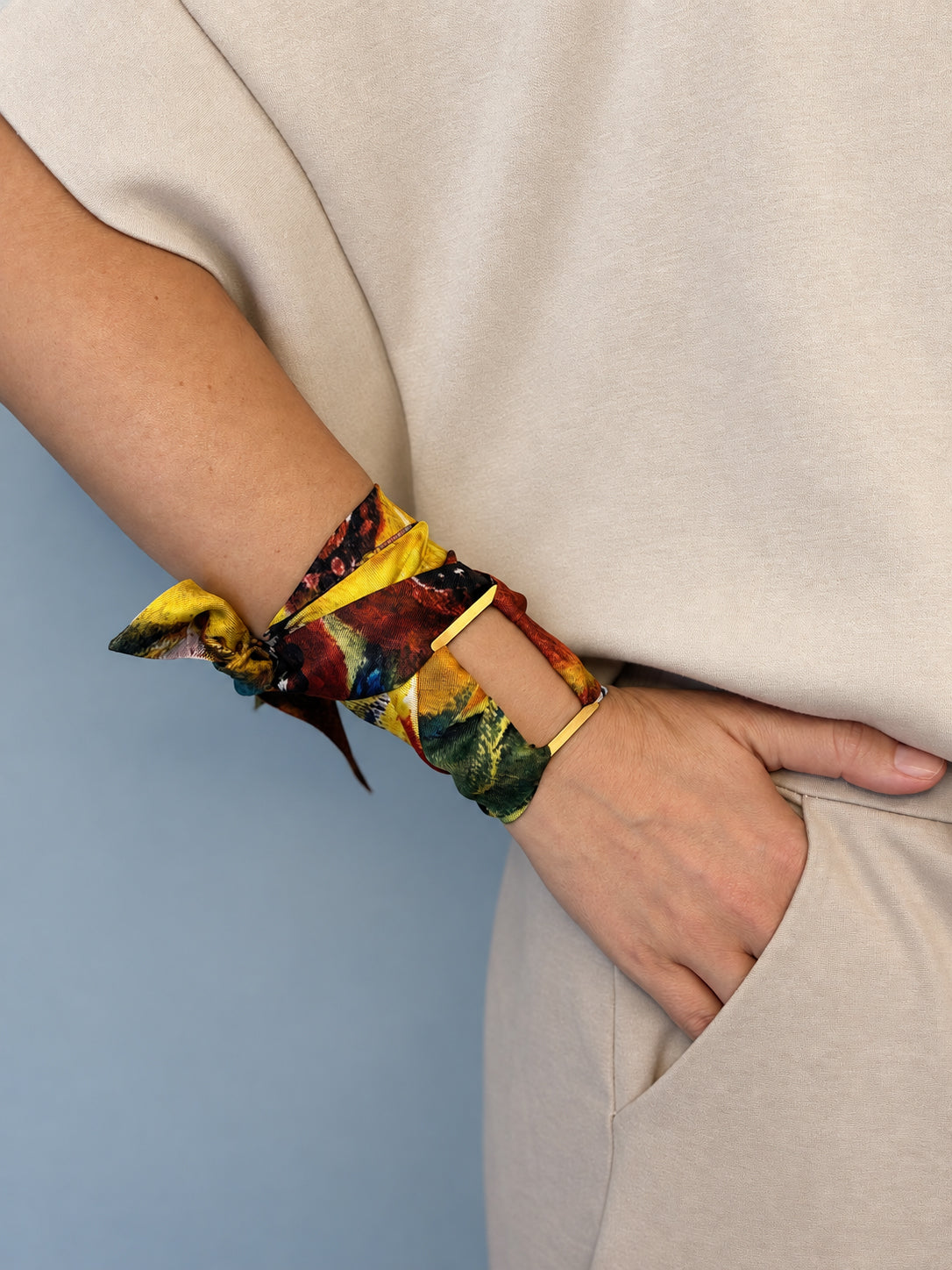 A close-up of a person's wrist wearing a colorful, patterned fabric wrist wrap tied in a knot, paired with a thin gold bangle, set against a neutral beige top and a soft blue background.