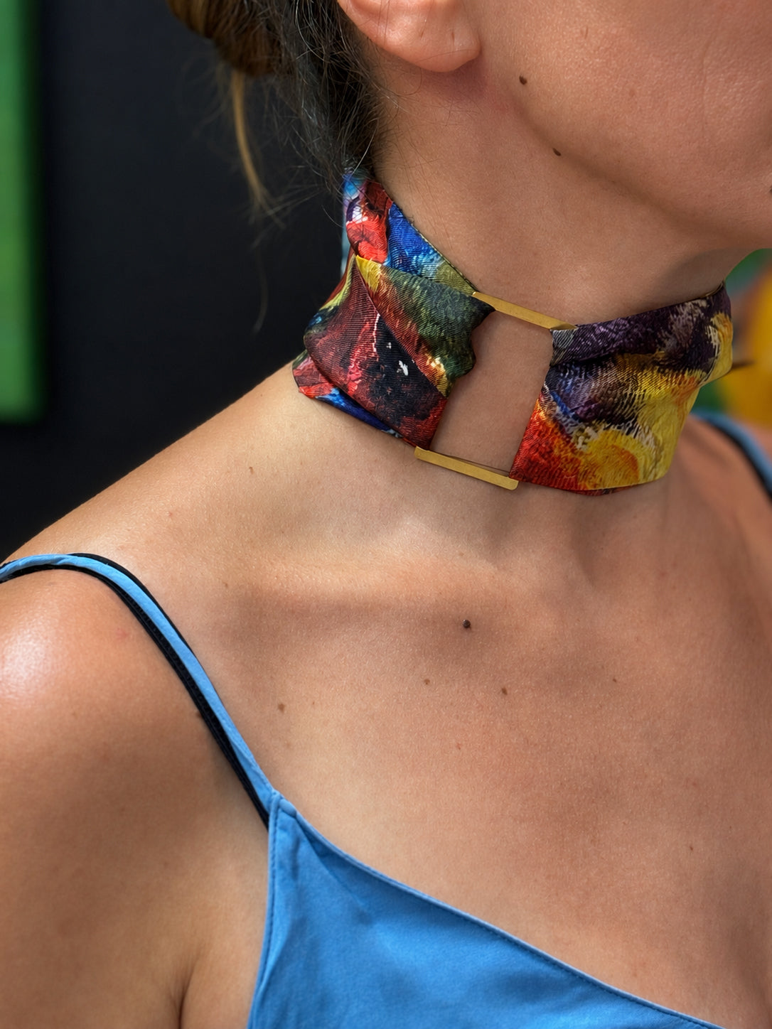 A close-up of a woman's neck and upper chest wearing a colorful abstract-patterned choker necklace with a cutout at the center and thin gold-toned side bars.