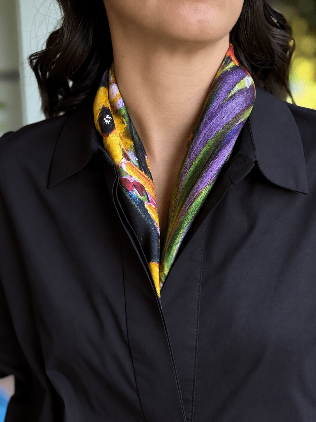 A close-up of a person wearing a black collared shirt and a colorful patterned silk scarf. The scarf features a vibrant design with purple, yellow, green, and pink tones.