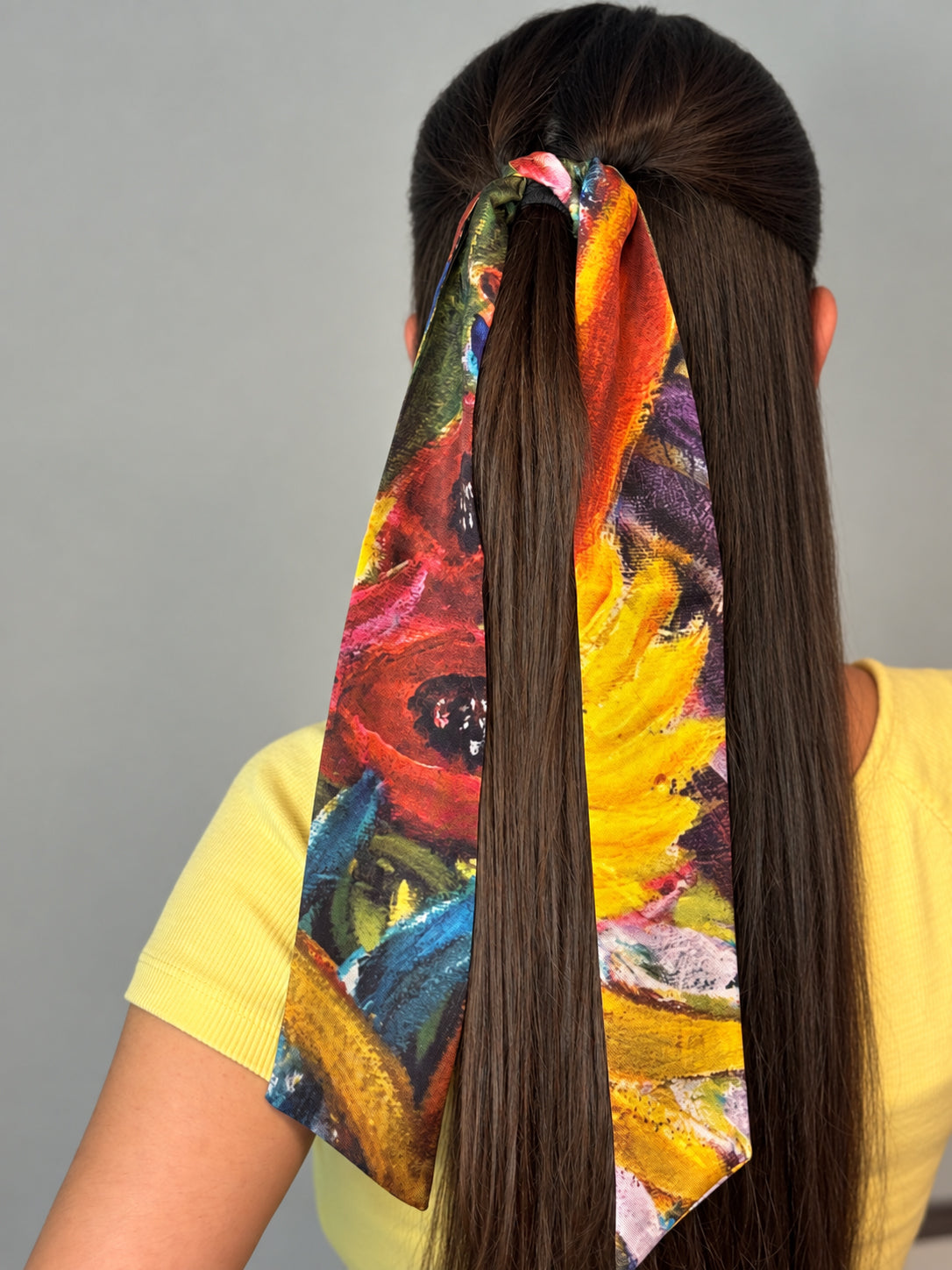 A long, dark brown hair accessory with a colorful abstract floral print tied around a high ponytail, shown on a model wearing a yellow shirt against a plain gray background.